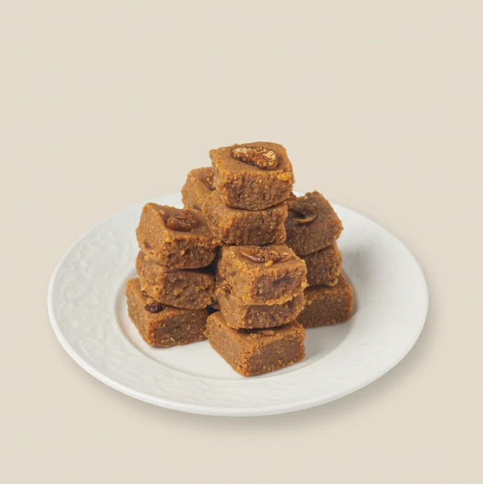 MM Mithaiwala Anjeer Cake Barfi with visible fig pieces, stacked on a white patterned plate against a beige background.