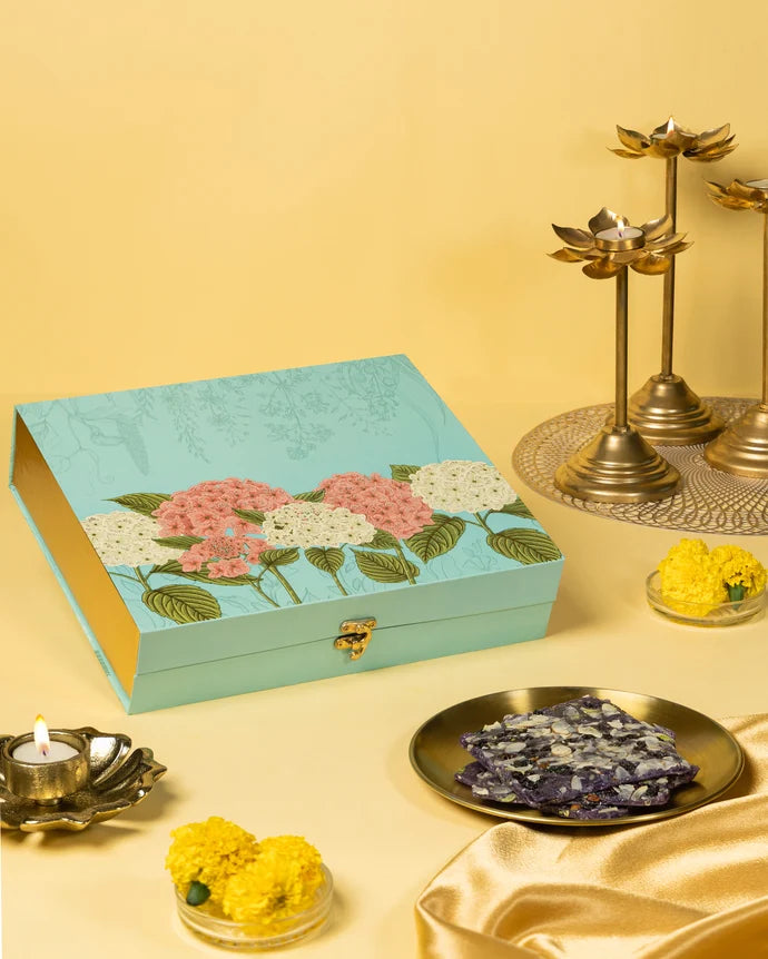 MM Mithaiwala Blackberry Ice Halwa Gift Box with a turquoise floral lid, displayed with halwa, marigolds and brass lamps.