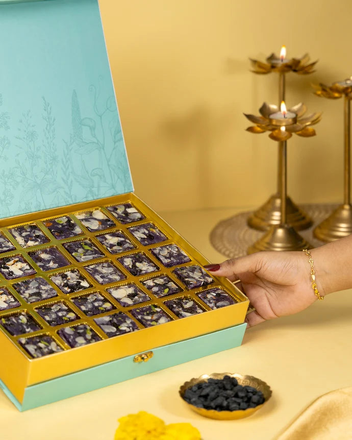 MM Mithaiwala Blackberry Ice Halwa Gift Box held open to show purple nut-garnished halwa in gold compartments, with brass lamps behind.