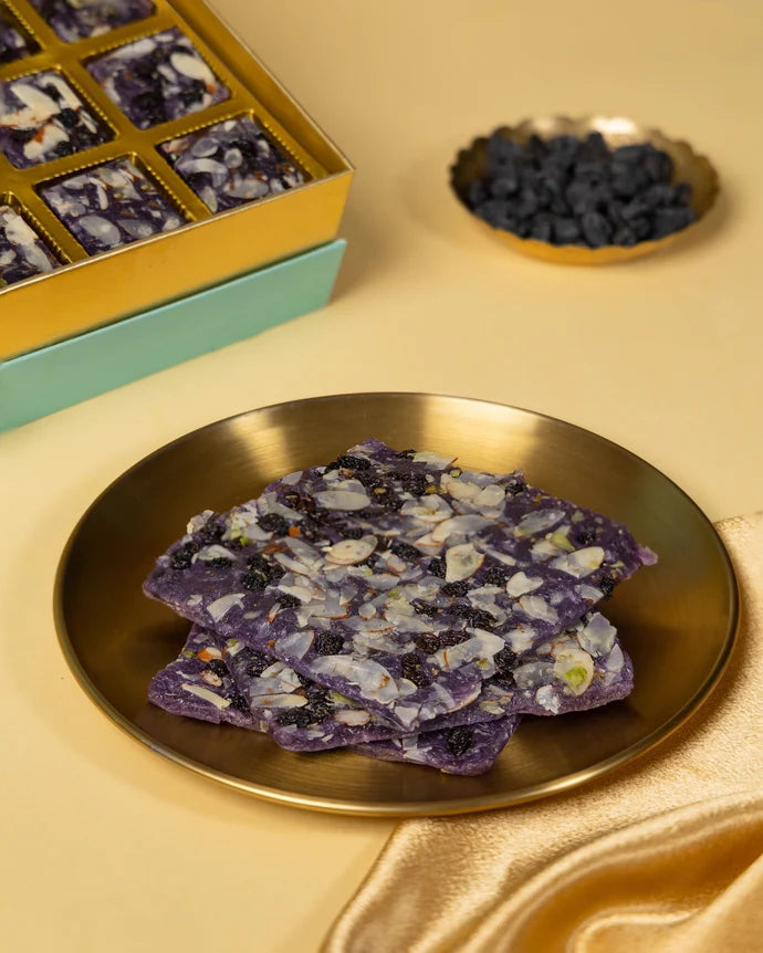 MM Mithaiwala Blackberry Ice Halwa Gift Box opened to display purple halwa topped with nuts and dried fruit in gold compartments beside brass lamps.