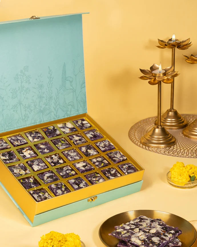 MM Mithaiwala Blackberry Ice Halwa Gift Box opened to display purple halwa topped with nuts and dried fruit in gold compartments beside brass lamps.