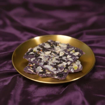 MM Mithaiwala purple Blackberry Ice Halwa garnished with sliced nuts and dried fruit on a gold plate over purple fabric.