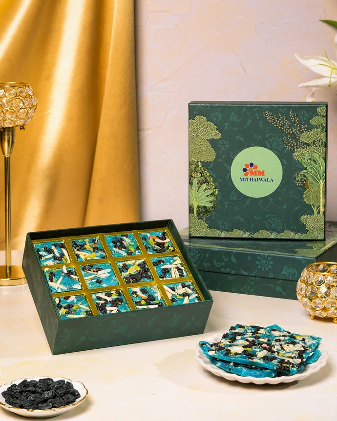 MM Mithaiwala Blueberry Ice Halwa Gift Box with a green botanical lid and twelve gold compartments, displayed with halwa and festive candle holders.