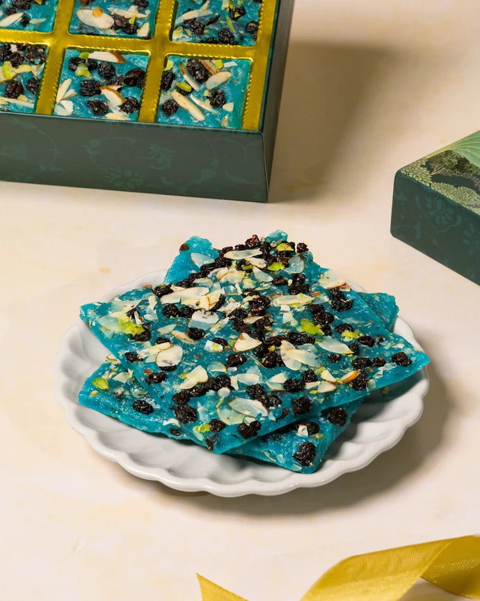 MM Mithaiwala Blueberry Ice Halwa topped with sliced nuts and dried fruit on a white scalloped plate, with the gift box behind.