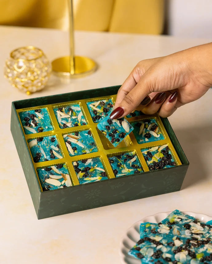 MM Mithaiwala Blueberry Ice Halwa Gift Box with a hand lifting a piece from its gold compartments.