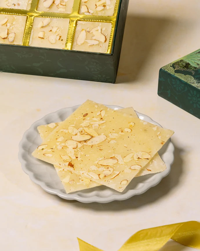 MM Mithaiwala Bombay Ice Halwa White Gift Box with a green botanical lid and twelve gold compartments, displayed with halwa and festive candle holders.