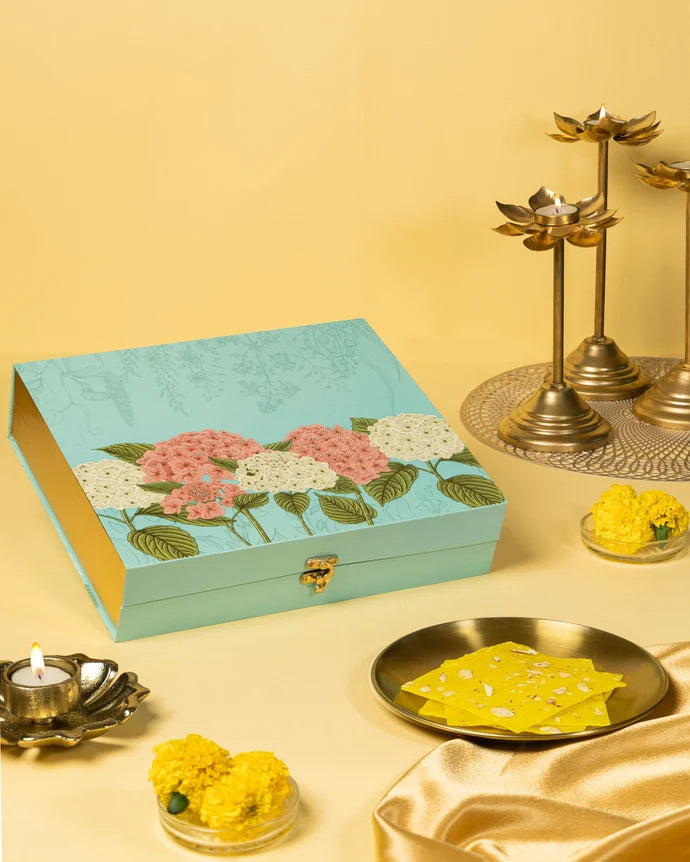 MM Mithaiwala Bombay Ice Halwa Yellow Gift Box with a turquoise floral lid, displayed with halwa, marigolds and brass lamps.