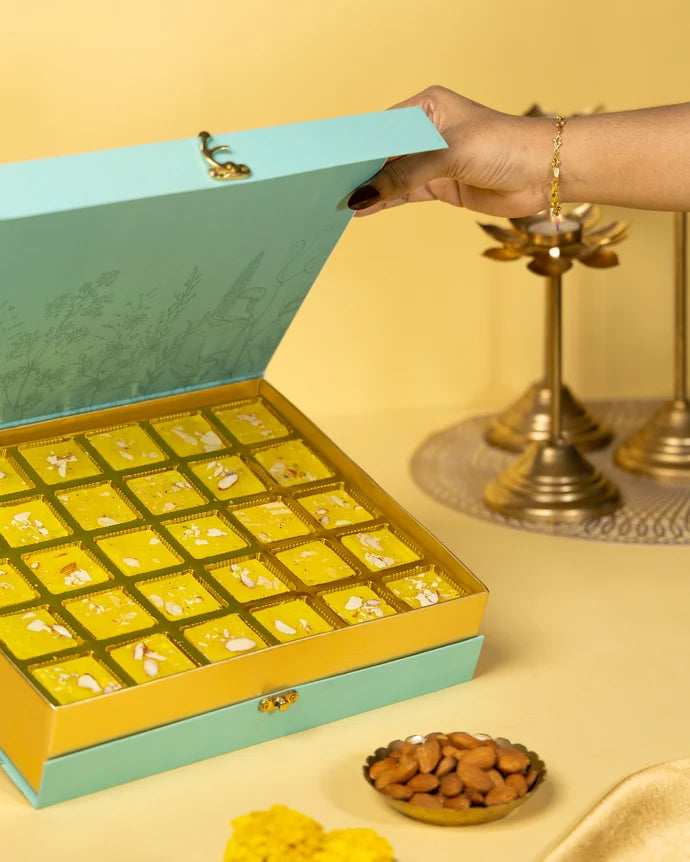 MM Mithaiwala Bombay Ice Halwa Yellow Gift Box with a hand lifting the lid to reveal halwa in gold compartments.