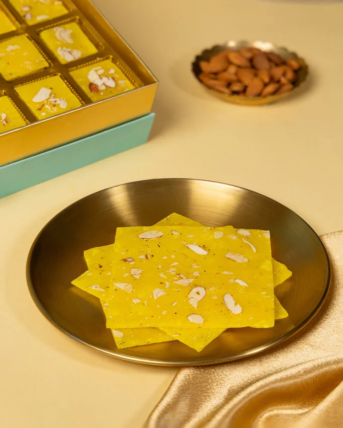 MM Mithaiwala Bombay Ice Halwa Yellow Gift Box opened to display almond-garnished halwa in gold compartments beside brass lamps.