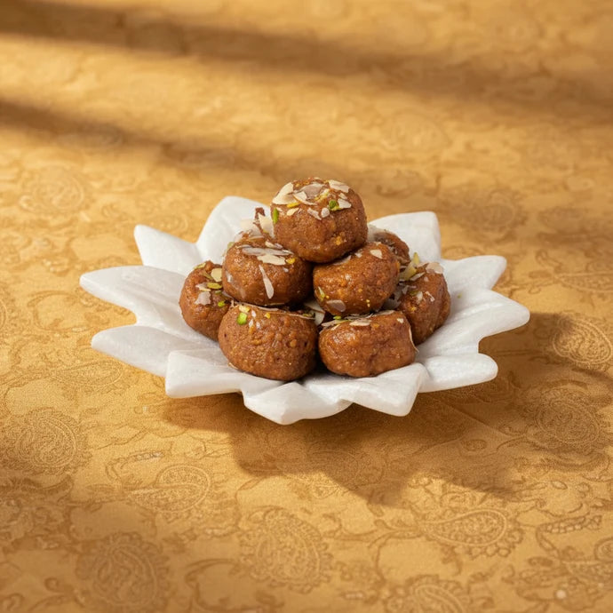 MM Mithaiwala Cake Peda topped with almond and pistachio slivers, stacked on a white flower-shaped plate against a golden background.