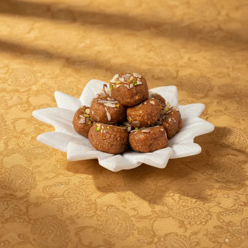MM Mithaiwala Cake Peda topped with almond and pistachio slivers, stacked on a white flower-shaped plate against a golden background.