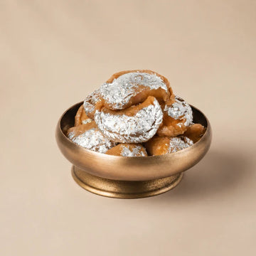 MM Mithaiwala Chandrakala (Gunjiya) topped with silver leaf in a brass bowl on a beige background.