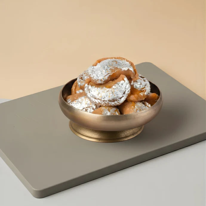 MM Mithaiwala Chandrakala (Gunjiya) topped with silver leaf in a brass bowl on a beige background.