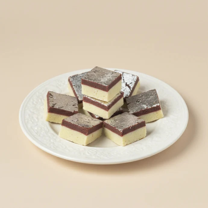 MM Mithaiwala Chocolate Barfi with cream and chocolate layers topped with silver leaf, stacked on a white patterned plate.