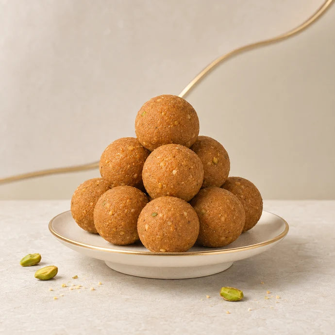 MM Mithaiwala golden-brown Churma Laddu stacked on a white gold-rimmed plate with pistachios alongside.