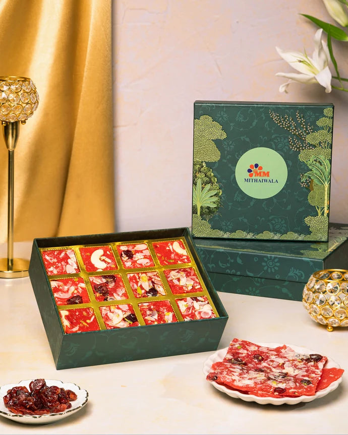 MM Mithaiwala Cranberry Ice Halwa Gift Box with a green botanical lid and twelve gold compartments, displayed with halwa and festive candle holders.