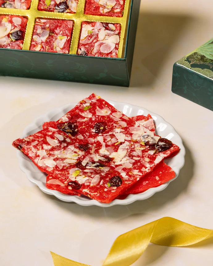 MM Mithaiwala Cranberry Ice Halwa Gift Box with a green botanical lid and twelve gold compartments, displayed with halwa and festive candle holders.