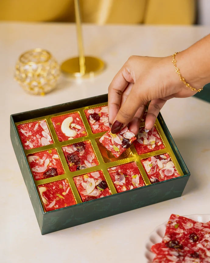 MM Mithaiwala Cranberry Ice Halwa Gift Box with a hand lifting a nut-garnished piece from its gold compartments.