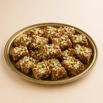 MM Mithaiwala Dry Fruit Barfi squares topped with chopped nuts, arranged on a brass tray against a beige background.
