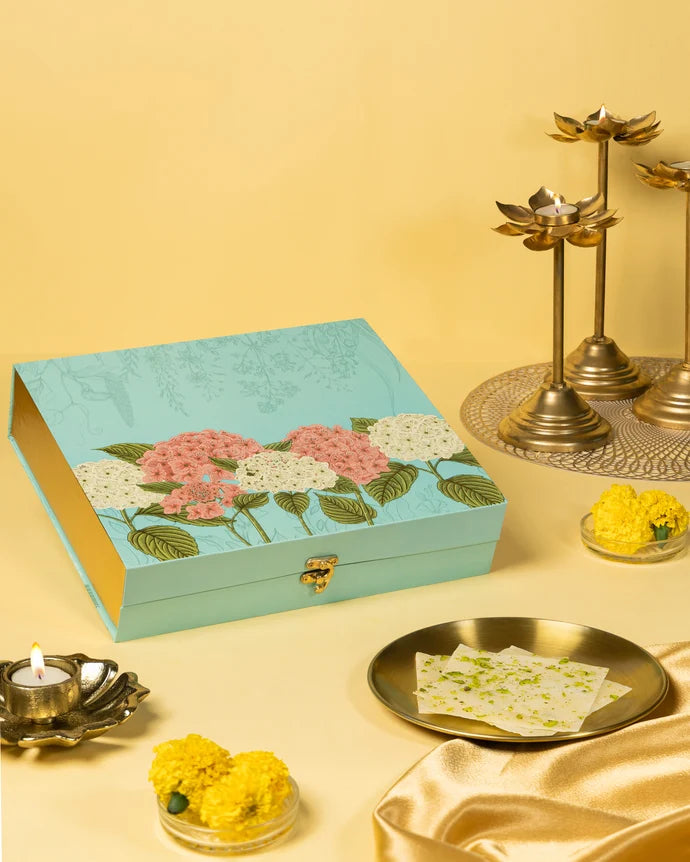 MM Mithaiwala Golden Ice Halwa Gift Box with a turquoise floral lid, displayed with halwa, marigolds and brass lamps.