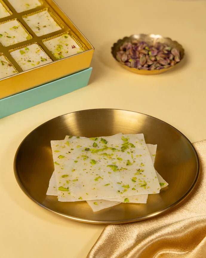 MM Mithaiwala Golden Ice Halwa Gift Box opened to display pistachio-garnished halwa in gold compartments beside brass lamps.