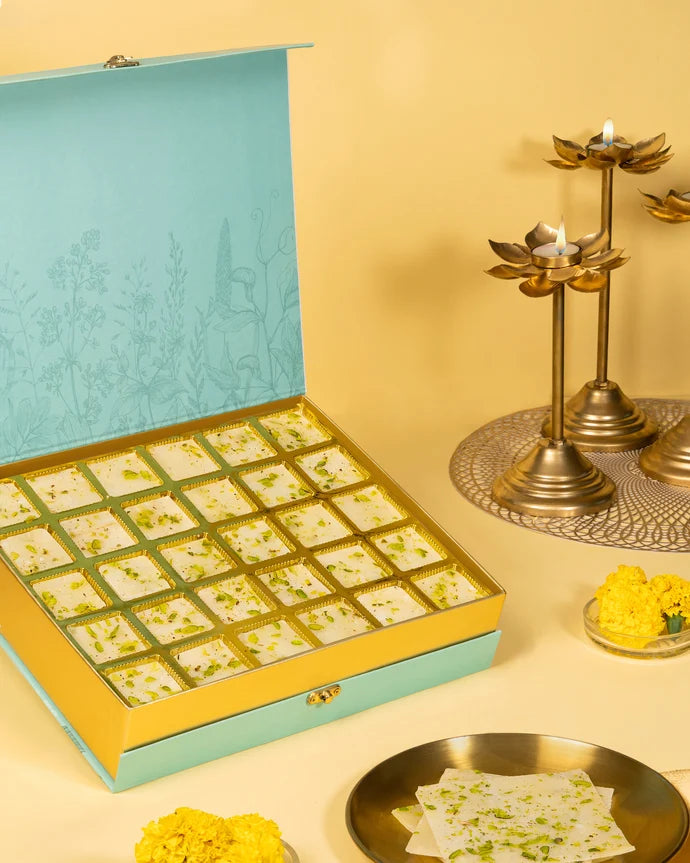MM Mithaiwala Golden Ice Halwa Gift Box opened to display pistachio-garnished halwa in gold compartments beside brass lamps.