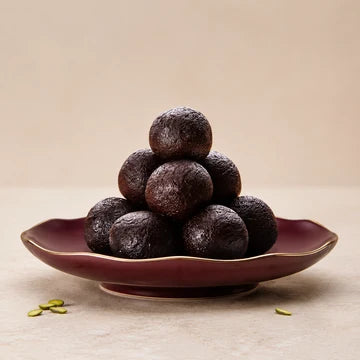 MM Mithaiwala Kala Angoor, dark round sweets stacked on a maroon plate against a beige background.