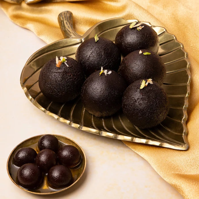 MM Mithaiwala Kala Jamun garnished with sliced nuts on a brass leaf-shaped platter, with a small bowl of kala jamun in syrup alongside.