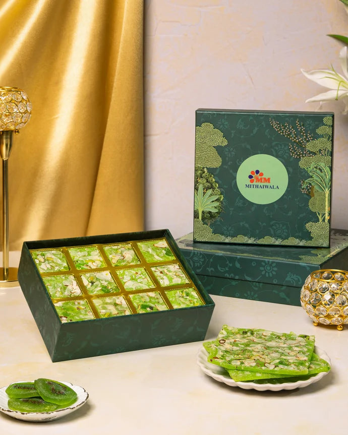 MM Mithaiwala Kiwi Ice Halwa Gift Box with a green botanical lid and twelve gold compartments, displayed with halwa and kiwi slices.