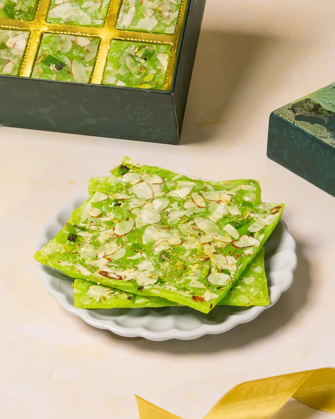 MM Mithaiwala Kiwi Ice Halwa Gift Box with a green botanical lid and twelve gold compartments, displayed with halwa and kiwi slices.