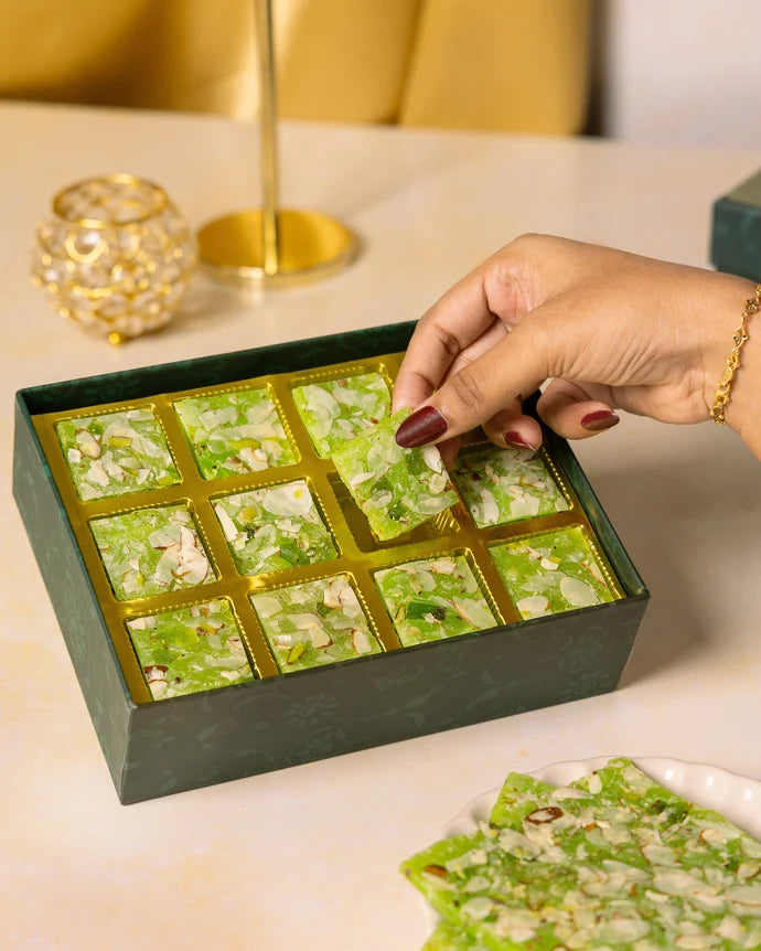 MM Mithaiwala Kiwi Ice Halwa Gift Box with a hand lifting a nut-garnished piece from its gold compartments.