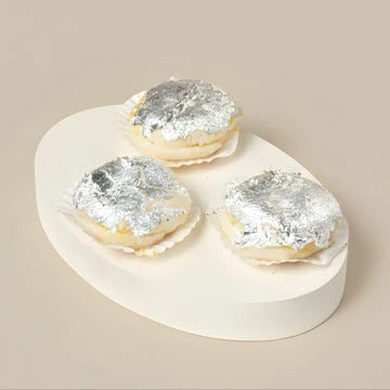 MM Mithaiwala Malai Chap topped with silver leaf and filled with cream, arranged on a white oval platter against a beige background.