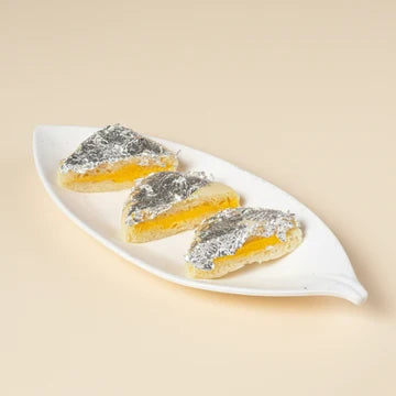 MM Mithaiwala triangular Malai Sandwich with yellow filling and silver leaf, arranged on a white leaf-shaped platter.