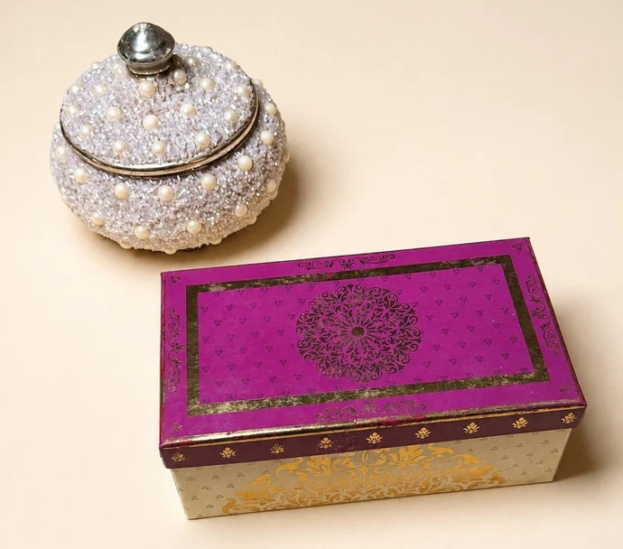 MM Mithaiwala Mithai Mood Box, a closed cream and gold gift box with a magenta patterned lid beside a decorative pearl-studded jar.