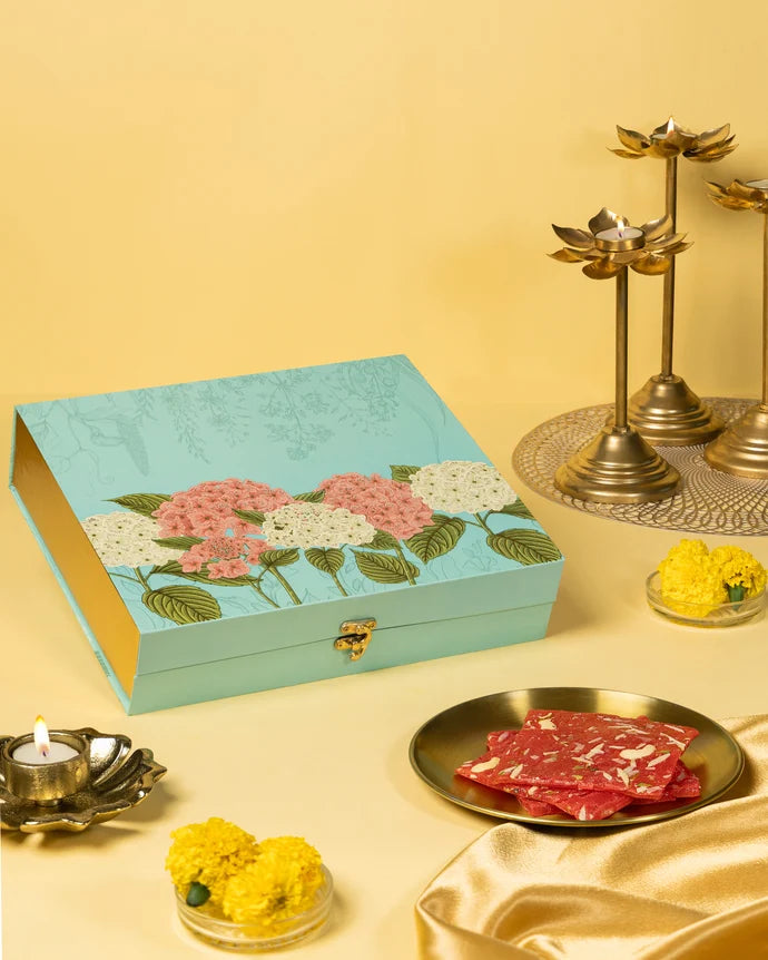 MM Mithaiwala Mohini Ice Halwa Gift Box with a turquoise floral lid, displayed with halwa, brass lamps and marigolds.