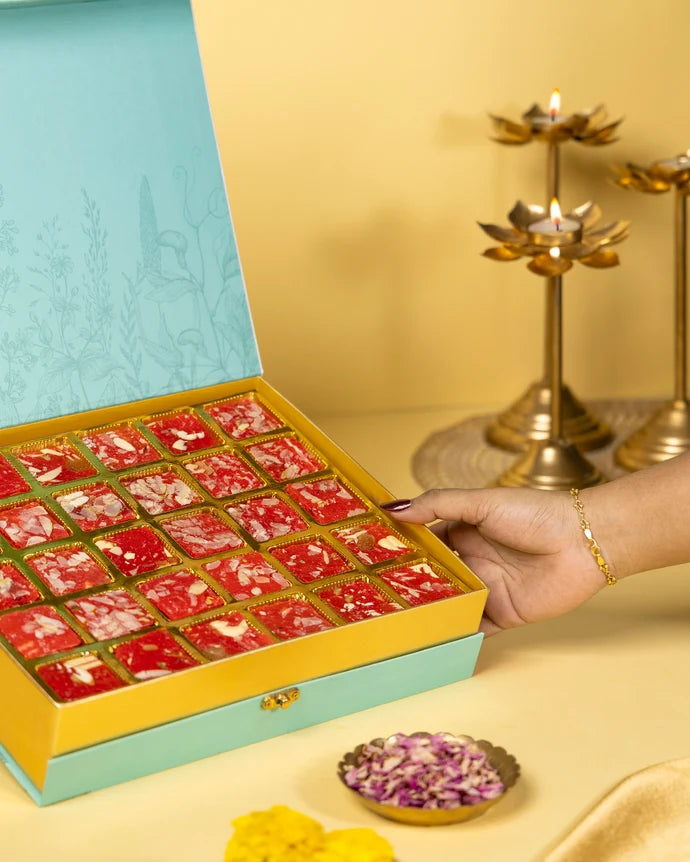MM Mithaiwala Mohini Ice Halwa Gift Box held open to show red halwa in gold compartments, with festive brass lamps behind.