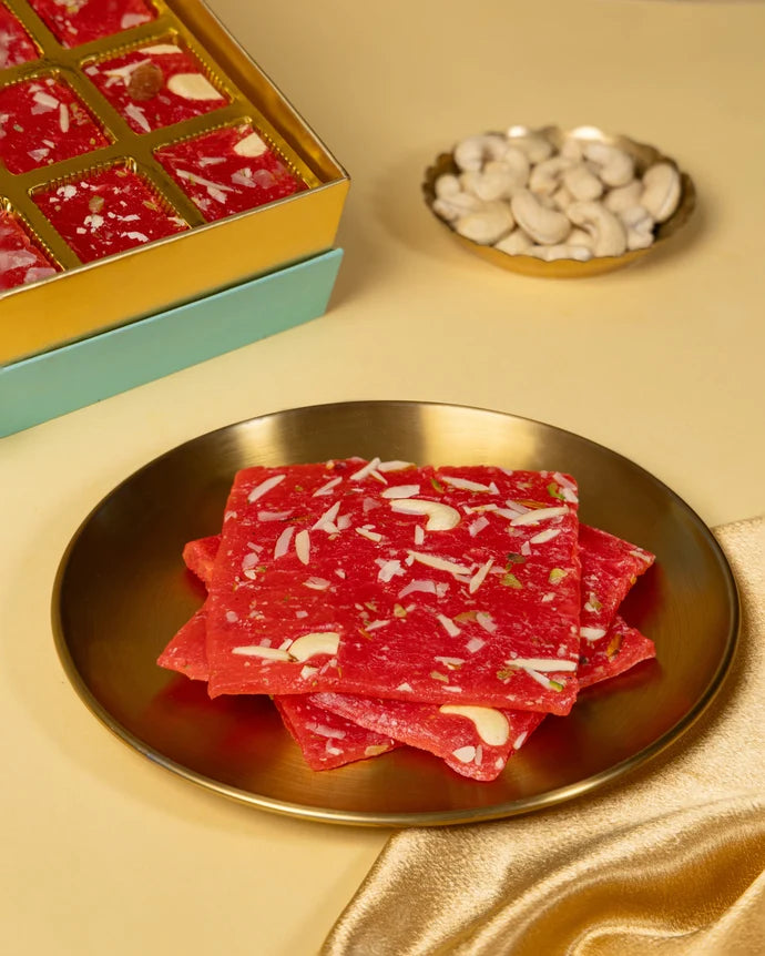MM Mithaiwala Mohini Ice Halwa topped with sliced nuts on a gold plate, with the open gift box behind.