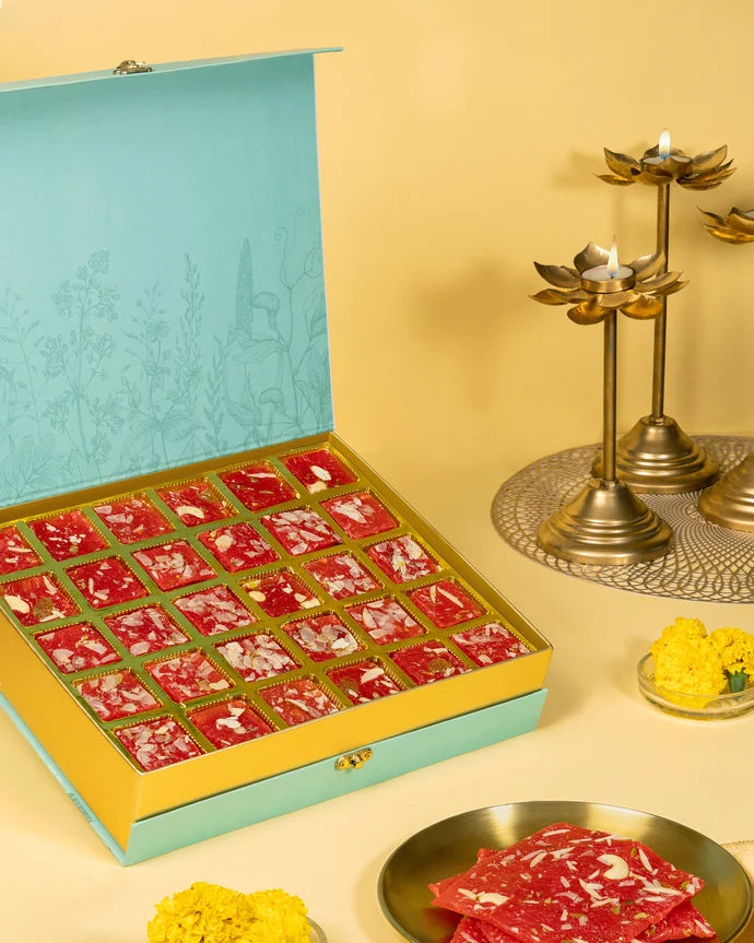 MM Mithaiwala Mohini Ice Halwa Gift Box opened to display red nut-garnished halwa in gold compartments, beside brass lamps.