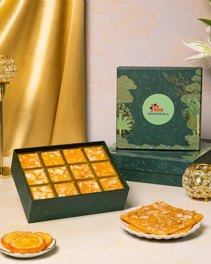 MM Mithaiwala Orange Ice Halwa Gift Box with a green botanical lid and twelve gold compartments, displayed with halwa and orange slices.