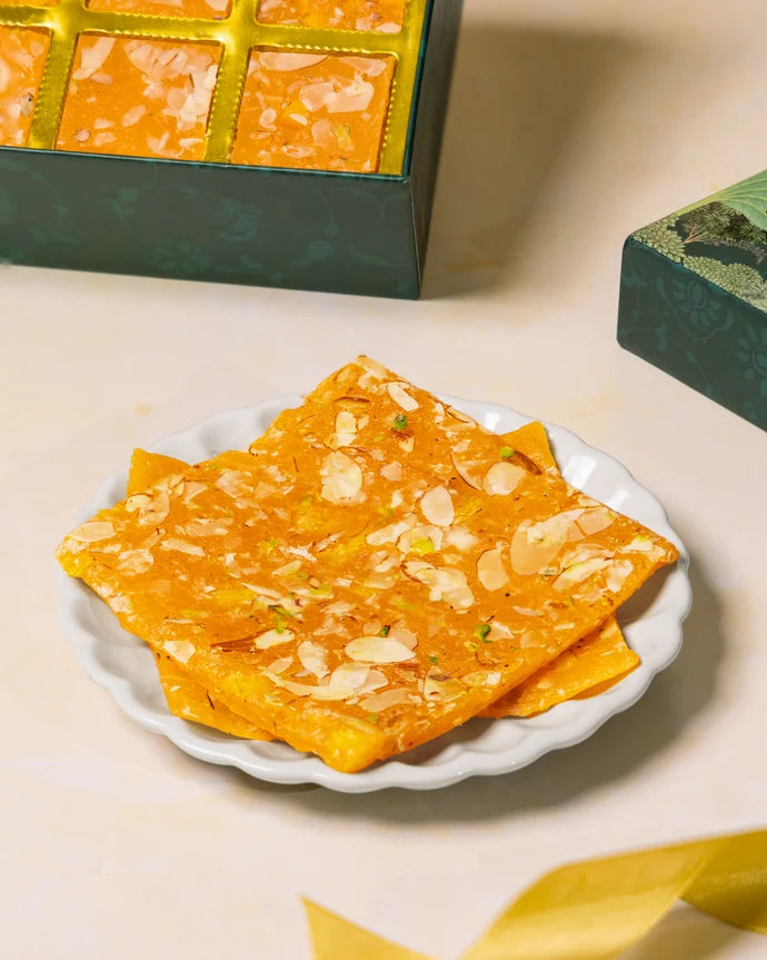 MM Mithaiwala Orange Ice Halwa topped with sliced almonds and pistachios on a white scalloped plate, with the gift box behind.