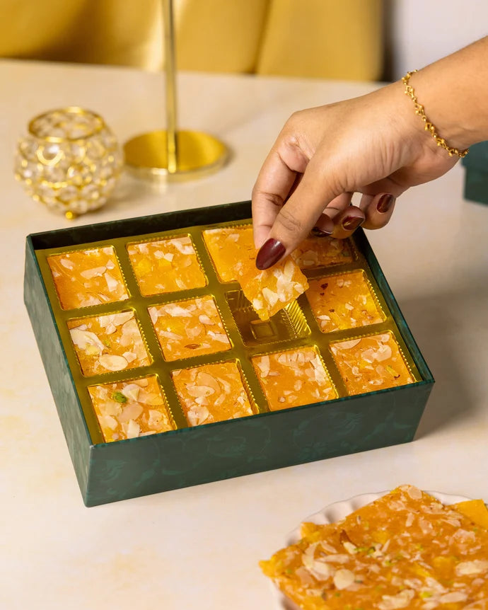 MM Mithaiwala Orange Ice Halwa Gift Box with a hand lifting an almond-garnished piece from its gold compartments.