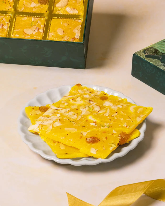 MM Mithaiwala yellow Pineapple Ice Halwa topped with sliced nuts and raisins on a white scalloped plate, with the gift box behind.