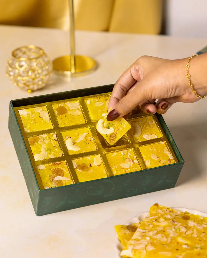 MM Mithaiwala Pineapple Ice Halwa Gift Box with a hand lifting a nut-garnished piece from its gold compartments.
