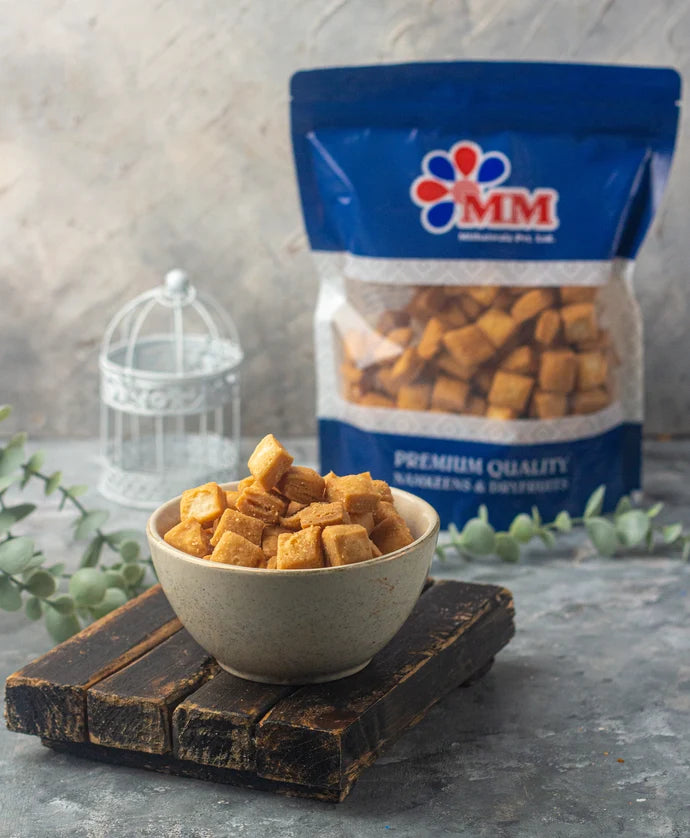 MM Mithaiwala Shakar Para Sweet, golden bite-sized squares served in a bowl on a wooden board with a branded blue pouch behind.