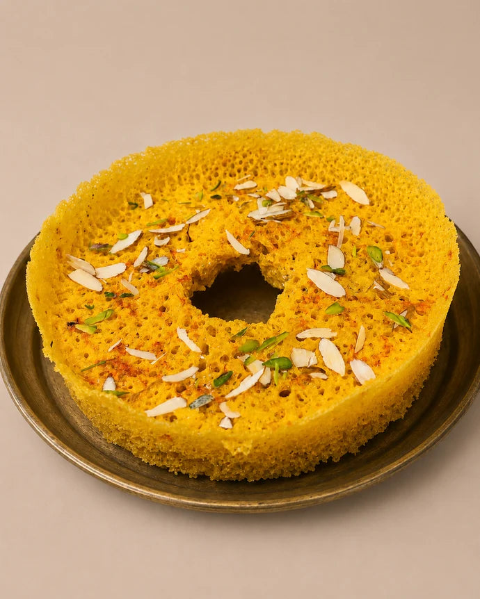 MM Mithaiwala Small Kesar Ghevar garnished with sliced nuts, with silver leaf on the centre piece, arranged on a maroon oval platter.