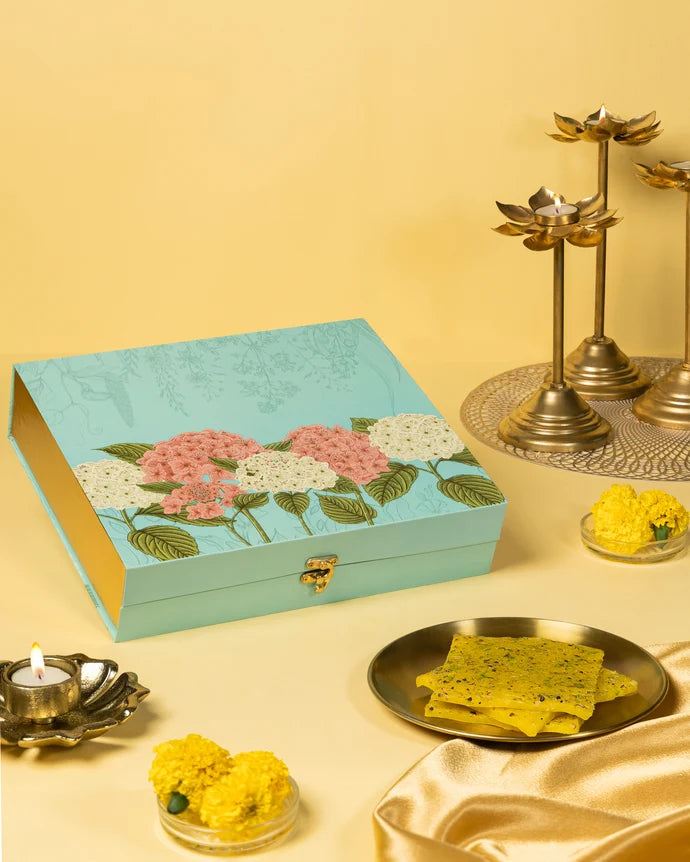 MM Mithaiwala Special Kesar Golden Ice Halwa Gift Box with a turquoise floral lid, displayed with halwa, marigolds and brass lamps.