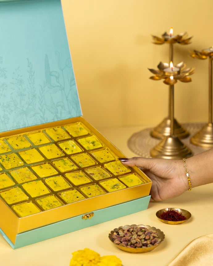MM Mithaiwala Special Kesar Golden Ice Halwa Gift Box held open to show yellow halwa in gold compartments, with saffron and pistachios alongside.