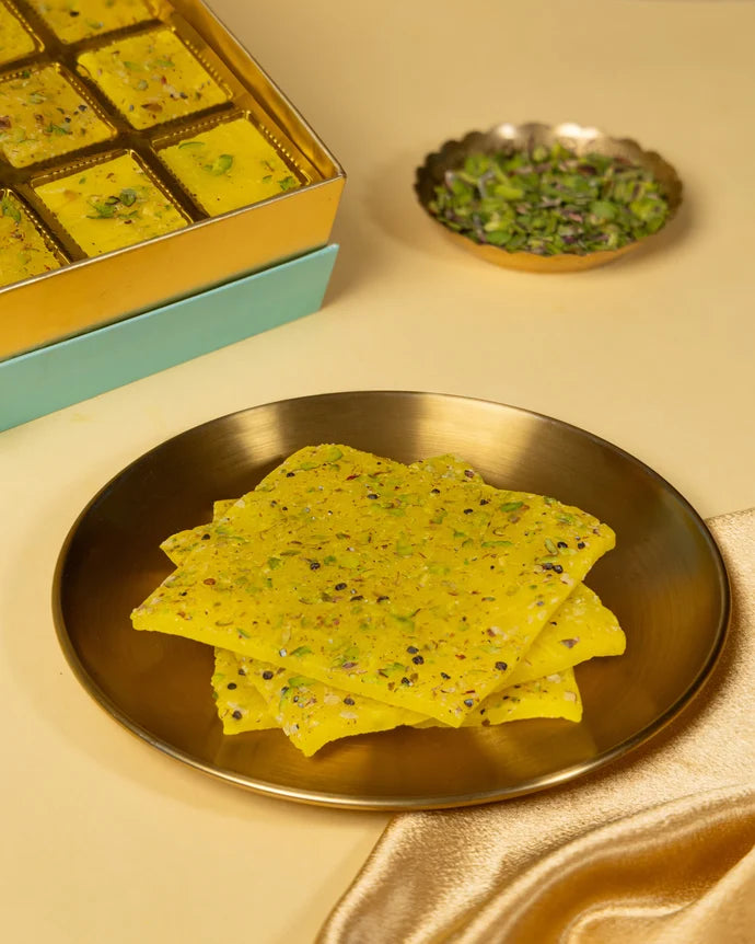 MM Mithaiwala Special Kesar Golden Ice Halwa topped with chopped pistachios on a gold plate, with the gift box behind.
