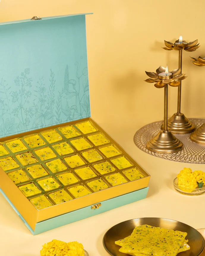 MM Mithaiwala Special Kesar Golden Ice Halwa Gift Box opened to display yellow pistachio-garnished halwa in gold compartments beside brass lamps.