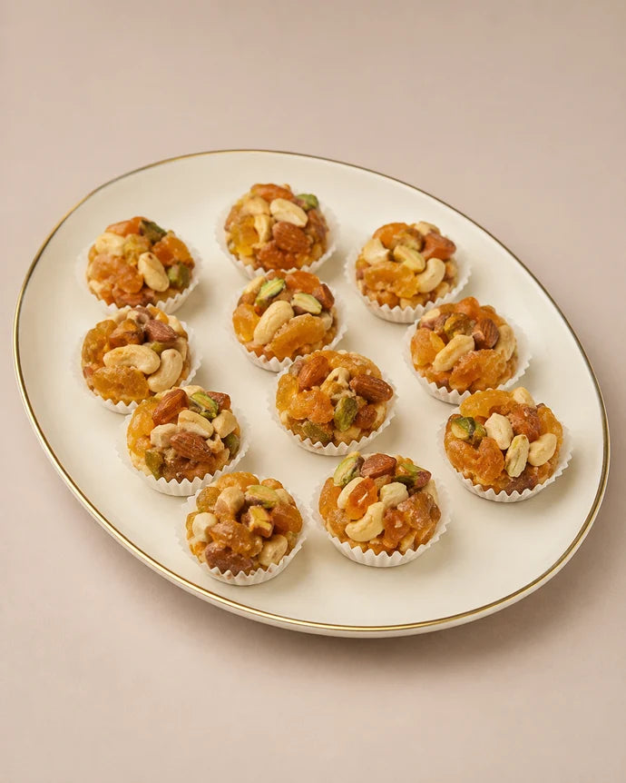 MM Mithaiwala Sugar Free Apricot Laddu with visible apricot pieces, almonds, cashews and pistachios, arranged in paper cups on a white gold-rimmed platter.